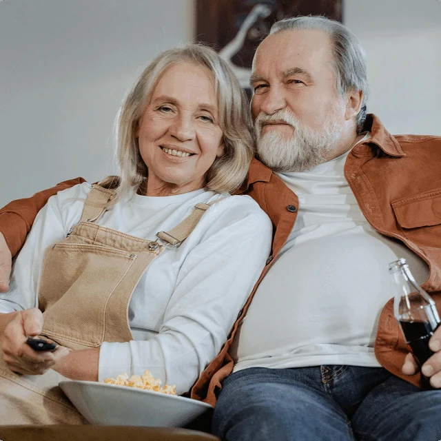 An older couple sits on a sofa together, smiling. One person holds a TV remote and a bowl of popcorn, while the other holds a glass bottle of soda.