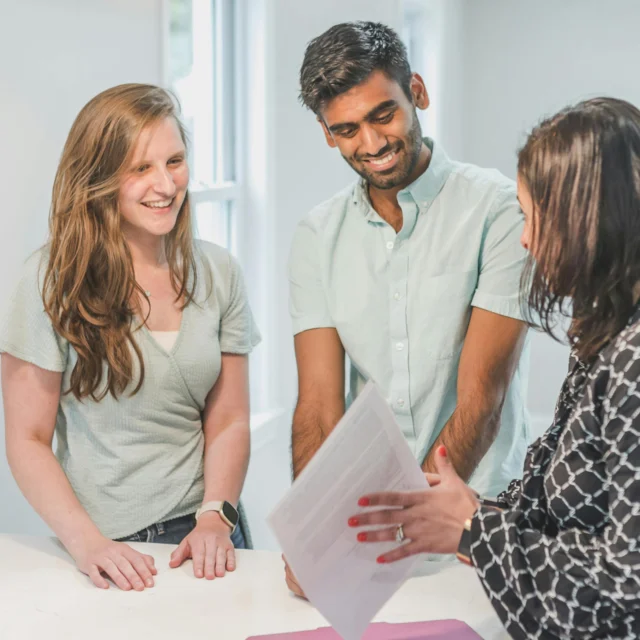 Three people stand together indoors; two listen and smile while the third person, holding a document, discusses its contents with them.