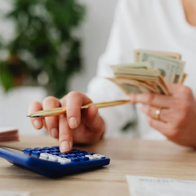 A person holds a stack of cash in one hand and uses a blue calculator with the other hand, seated at a table with papers.