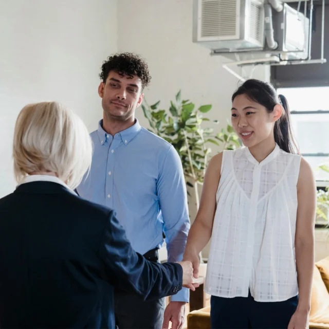 Three people in business attire stand indoors; a woman with blonde hair shakes hands with an Asian woman while a man stands next to them, watching. Plants and an air conditioner are visible in the background.