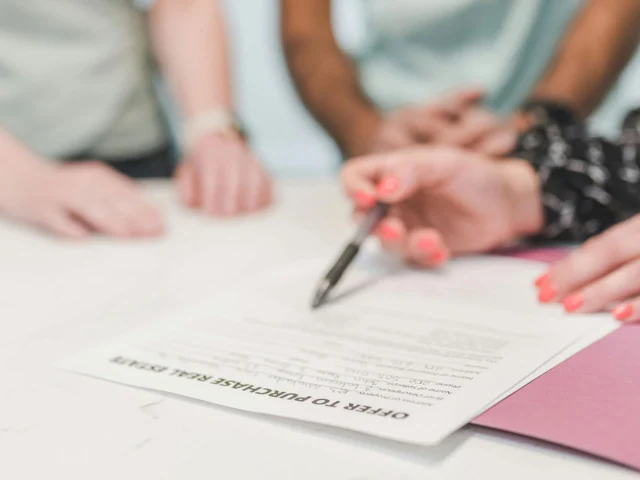 Three people review and sign an "Offer to Purchase Real Estate" document on a white table, with a visible pen and folder.