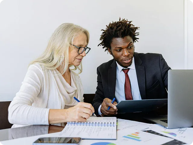 Two people, a woman and a man in business attire, sit at a desk reviewing documents and graphs with a laptop in front of them.