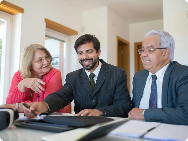 Three people in business attire sit at a table with documents, having a discussion in a bright office setting.