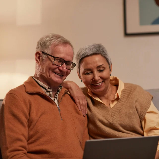 An older man and woman sit closely together on a couch, smiling as they look at a laptop screen.