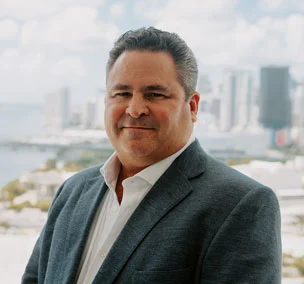 A middle-aged man in a gray suit jacket and white shirt stands outdoors with a blurred cityscape in the background.