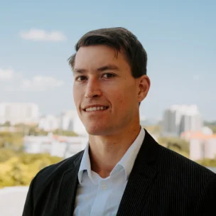 A man in a dark suit and white shirt stands outdoors with a cityscape and blue sky in the background.