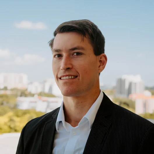 A man in a dark suit and white shirt stands outdoors with a cityscape and blue sky in the background.