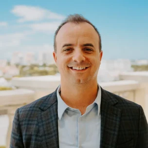 A man in a blazer and light blue shirt smiles while standing on a balcony with a cityscape and blue sky in the background.