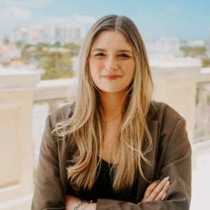 A woman with long blonde hair and a brown blazer stands on a balcony with buildings and trees visible in the background on a sunny day.