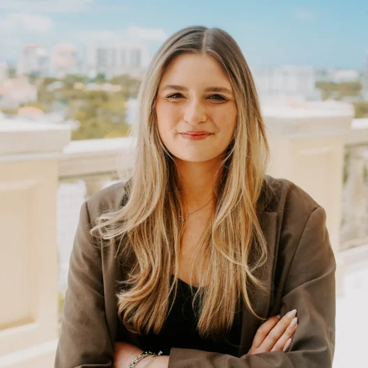 A woman with long blonde hair and a brown blazer stands on a balcony with buildings and trees visible in the background on a sunny day.