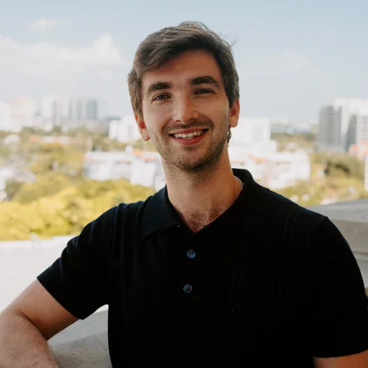A man with brown hair and a short beard, wearing a black polo shirt, stands on a balcony with a cityscape and trees in the background.